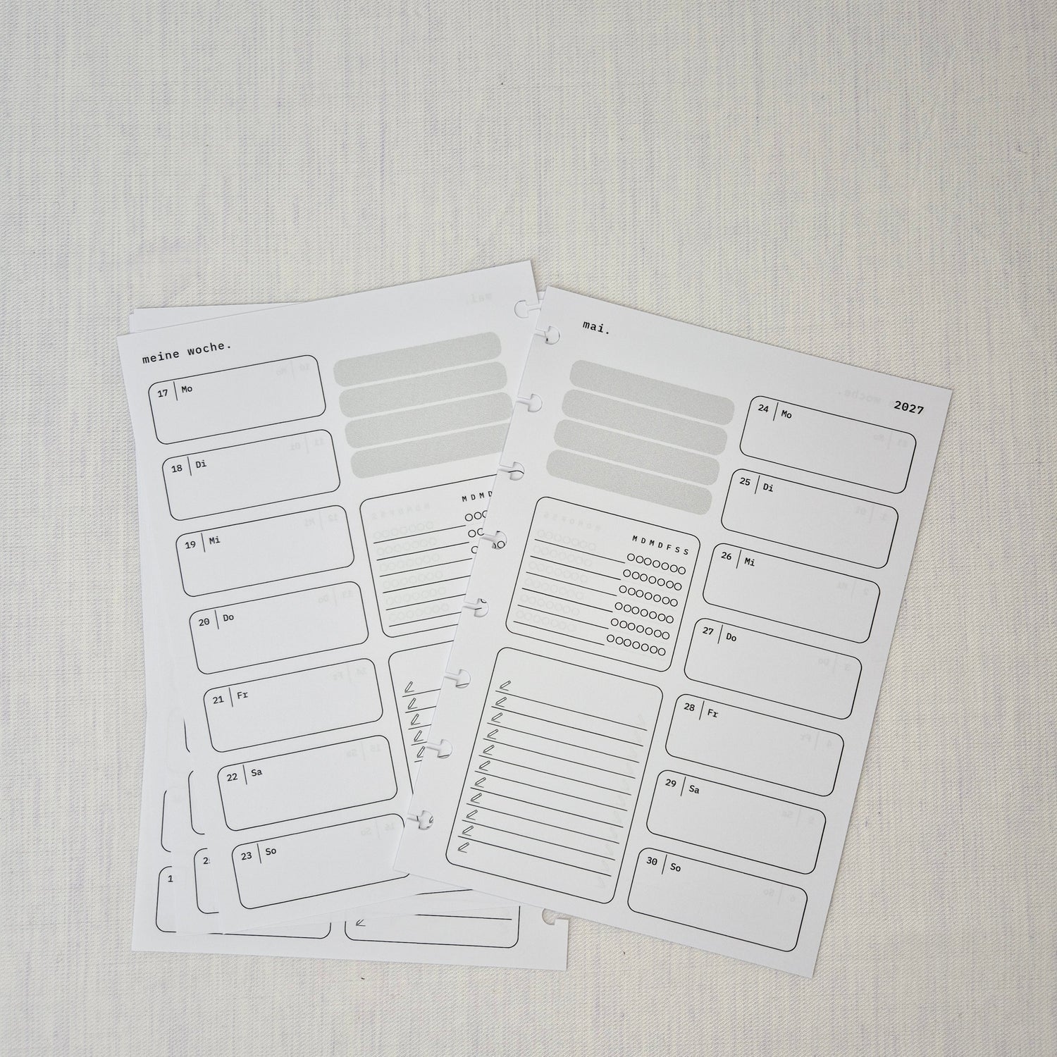 Zwei nebeneinanderliegende Planer-Einlagenseiten im minimalistischen Grau-Beige-Design mit der Überschrift WOCHE AUF 1 SEITE. Links eine Wochenübersicht ‘meine woche.’ mit Boxen für Mo–So und kleinen Datumsfeldern; rechts die Fortsetzung derselben Woche mit Boxen für Mo–So und dem Monatskopf ‘januar’ sowie Jahreszahl ‘2027’. In beiden Seiten gibt es zusätzlich neutrale Platzhalterfelder, einen kleinen Habit-Tracker mit Kreisen und einen linierten Notiz-/To-do-Bereich. Am Rand sind Lochungen/Discs-Markierung
