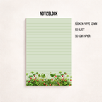 Liniertes Notizpapier im Hochformat mit grün-rosa Erdbeerwiesen-Motiv am unteren Rand, fotografiert auf dunkelgrauem Hintergrund. Links daneben liegt ein transparenter Stift parallel zum Block. Cleanes, minimalistisches Flatlay mit viel freiem Raum