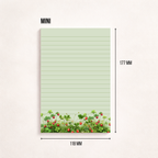 Liniertes Notizpapier im Hochformat mit grün-rosa Erdbeerwiesen-Motiv am unteren Rand, fotografiert auf dunkelgrauem Hintergrund. Links daneben liegt ein transparenter Stift parallel zum Block. Cleanes, minimalistisches Flatlay mit viel freiem Raum
