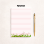 Minimalistischer, rosa linierter Notizblock mit Blumenwiesen-Motiv am unteren Rand auf hellem Hintergrund, rechts daneben ein goldener Kugelschreiber, darüber der Schriftzug „NOTIZBLOCK“