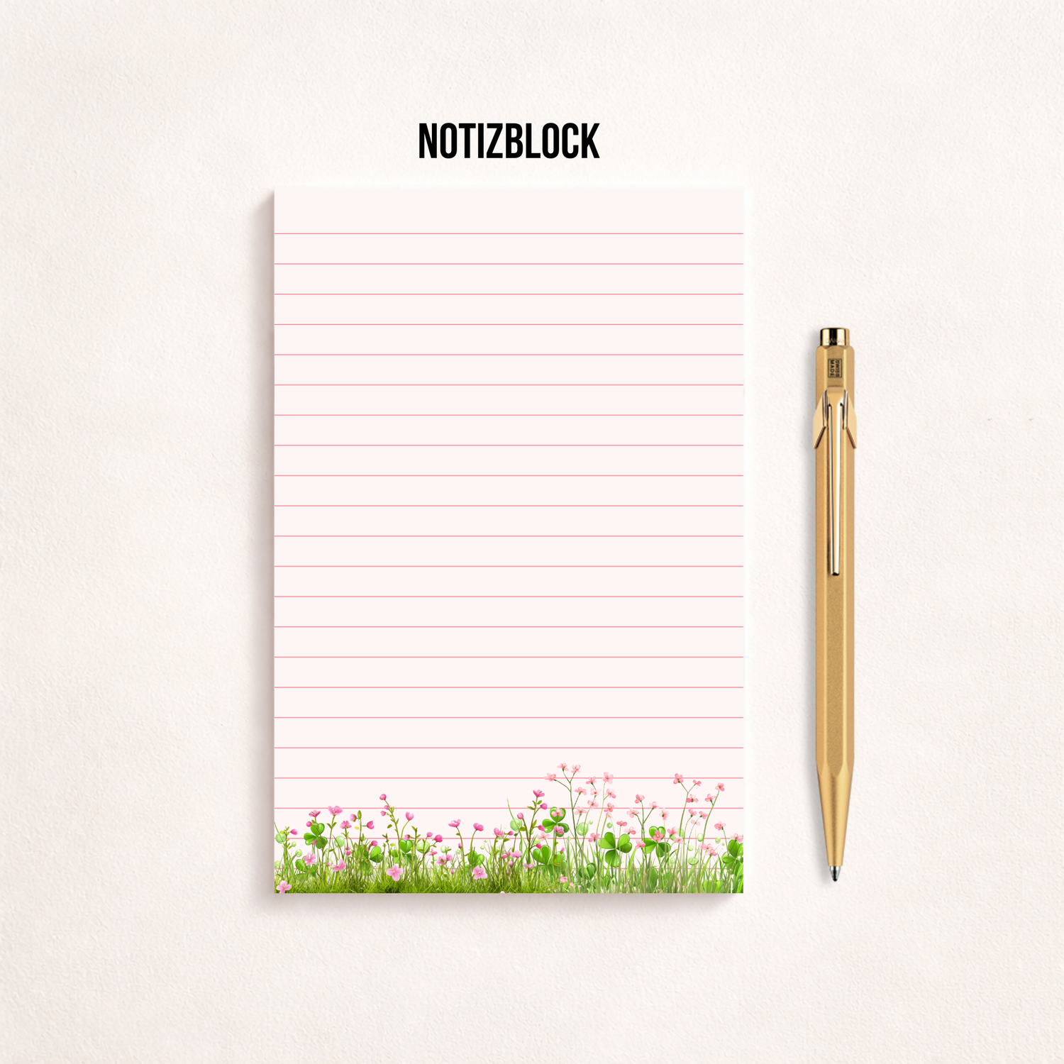 Minimalistischer, rosa linierter Notizblock mit Blumenwiesen-Motiv am unteren Rand auf hellem Hintergrund, rechts daneben ein goldener Kugelschreiber, darüber der Schriftzug „NOTIZBLOCK“