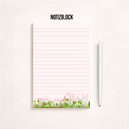 Minimalistischer, rosa linierter Notizblock mit Blumenwiesen-Motiv am unteren Rand auf hellem Hintergrund, rechts daneben ein goldener Kugelschreiber, darüber der Schriftzug „NOTIZBLOCK“